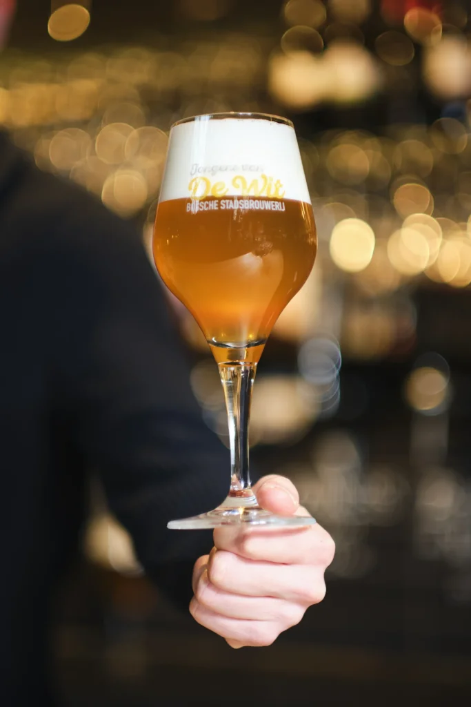 Hand houdt een glas troebel goudblond bier voor de verlichte bar