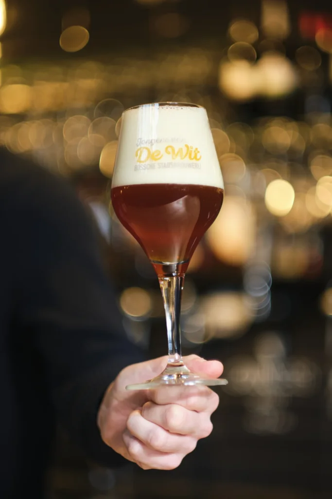 Hand houdt een glas amberkleurig bier met dikke schuimkraag voor de bar