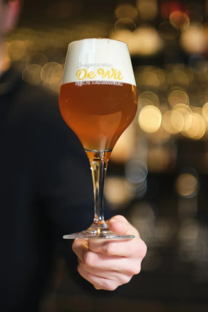 Hand houdt een glas amberblond bier in een De Wit-glas voor de bar
