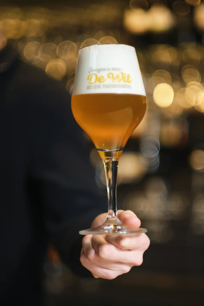 Hand houdt een glas lichtblond bier in een De Wit-glas voor de bar